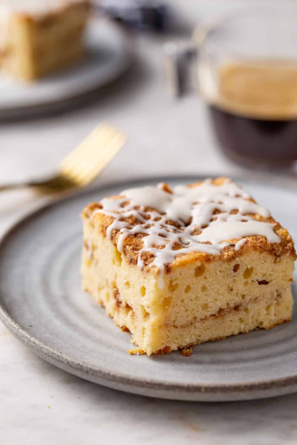 Angled view of cinnamon coffee cake on a white plate with gold fork and cup of espresso visible in the background. Cake topped with vanilla glaze