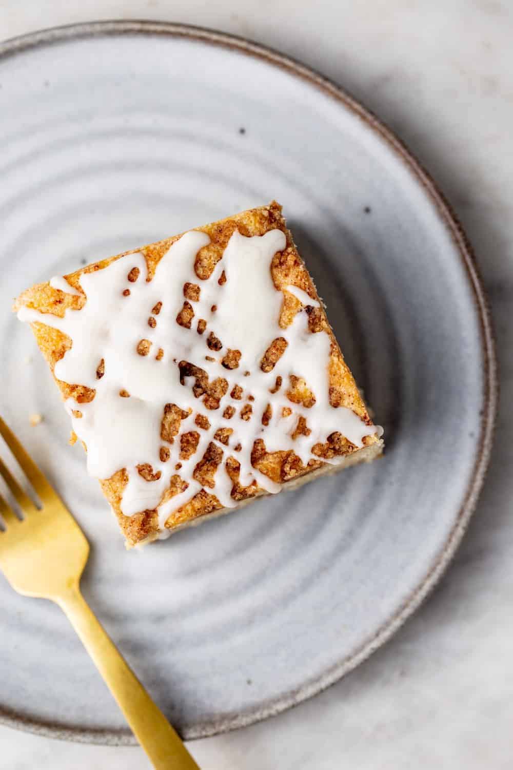 Overhead view of a slice of cinnamon coffee cake with vanilla glaze on a white plate next to a gold fork