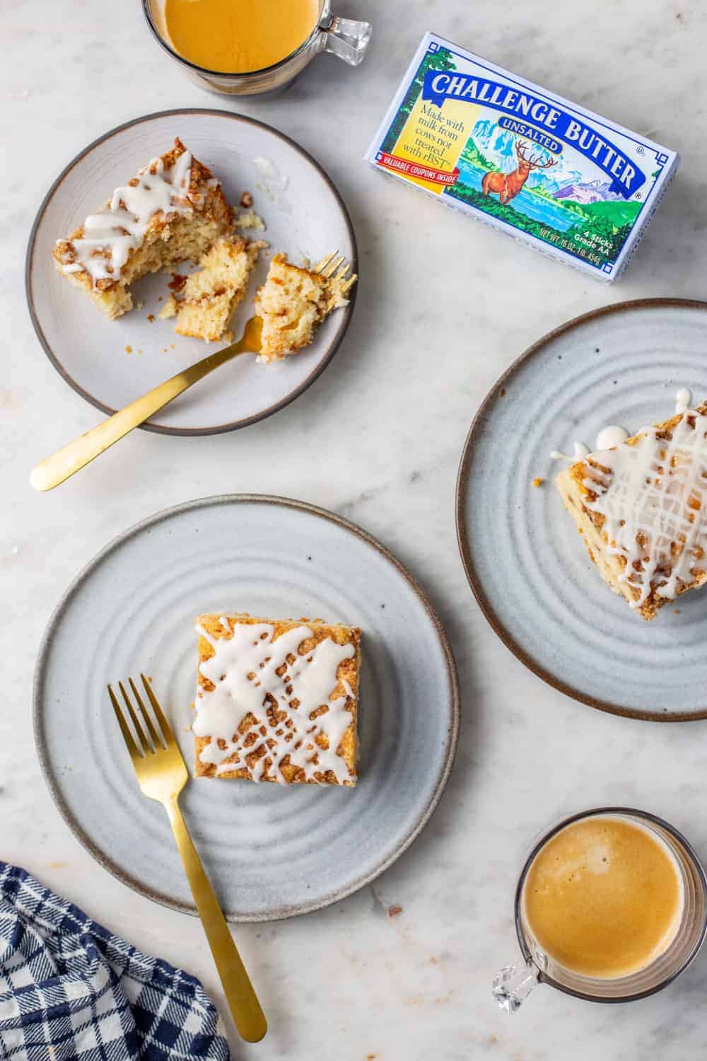 Three white plates with slices of cinnamon coffee cake arranged on a marble counter next to cups of coffee and a package of butter