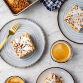 Cake pan with cinnamon coffee cake on a marble counter next to three plates of cinnamon coffee cake and two cups of espresso