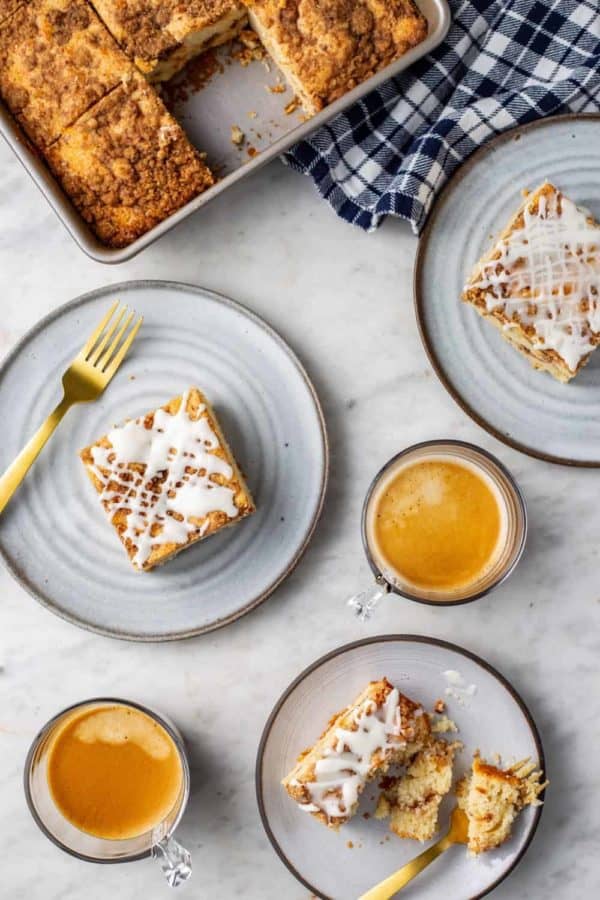 Cake pan with cinnamon coffee cake on a marble counter next to three plates of cinnamon coffee cake and two cups of espresso