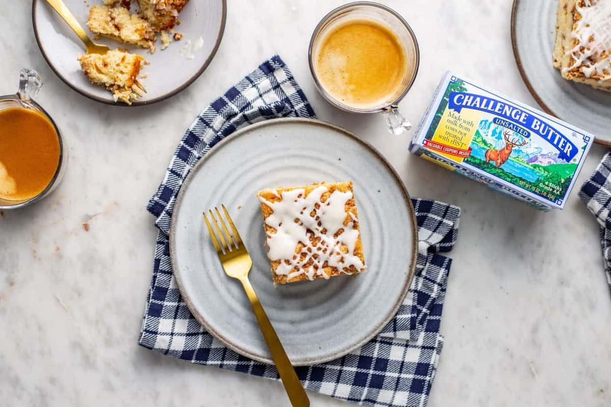 Overhead view of a slice of cinnamon coffee cake with vanilla glaze on a white plate sitting on a blue plaid napkin, next to a cup of espresso and a package of butter