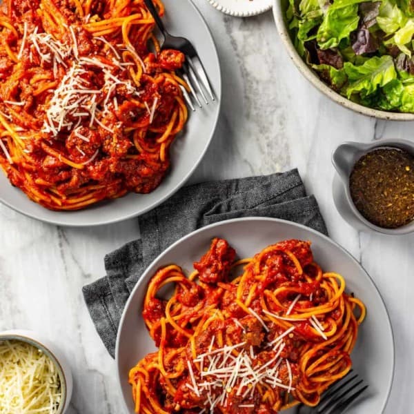 Spaghetti and homemade meat sauce portioned onto two gray plates on a marble surface, topped with parmesan cheese next to a green salad