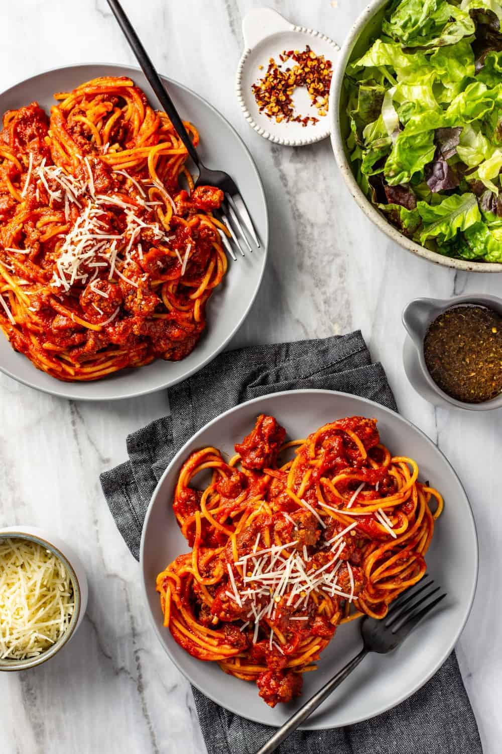 Spaghetti and homemade meat sauce portioned onto two gray plates on a marble surface, topped with parmesan cheese next to a green salad