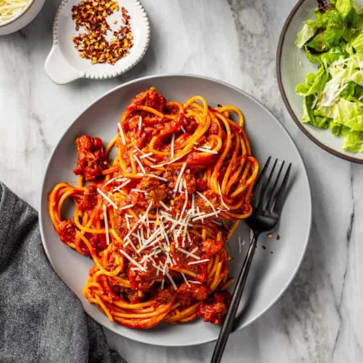 Spaghetti and homemade meat sauce on a gray plate with a fork, next to a green salad, all on a marble surface
