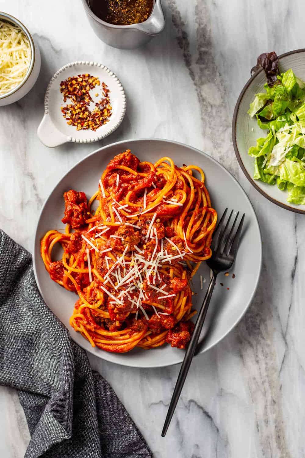 Spaghetti and homemade meat sauce on a gray plate with a fork, next to a green salad, all on a marble surface