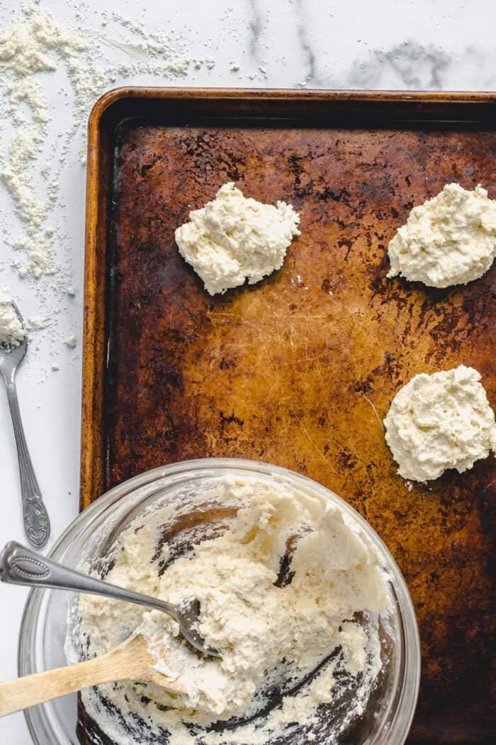 Drop biscuits being spooned onto a cookie sheet, with a spoon in the glass mixing bowl of biscuit dough