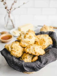 Basket lined with a dark gray napkin and filled with bisquick biscuits on a countertop with butter and honey