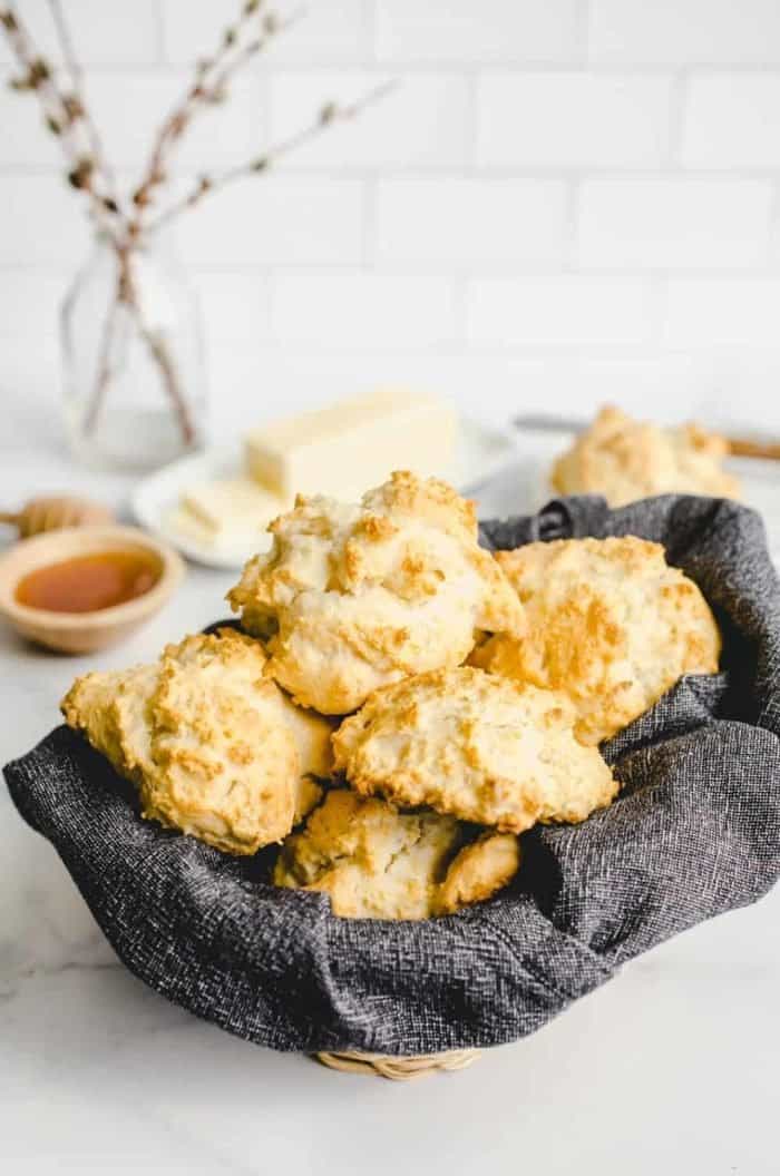Basket lined with a dark gray napkin and filled with bisquick biscuits on a countertop with butter and honey