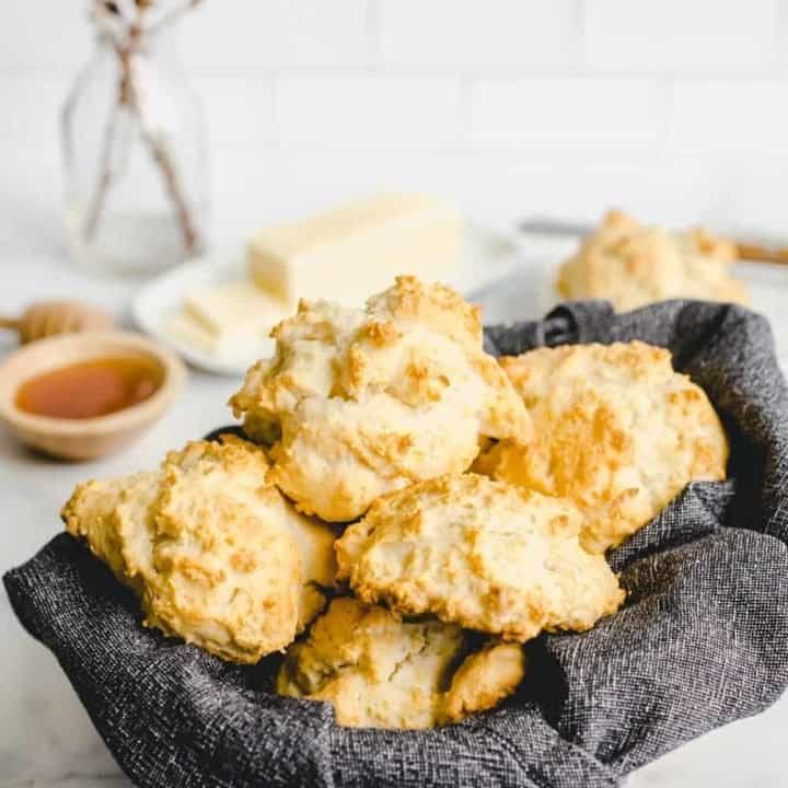 Basket lined with a dark gray napkin and filled with bisquick biscuits on a countertop with butter and honey