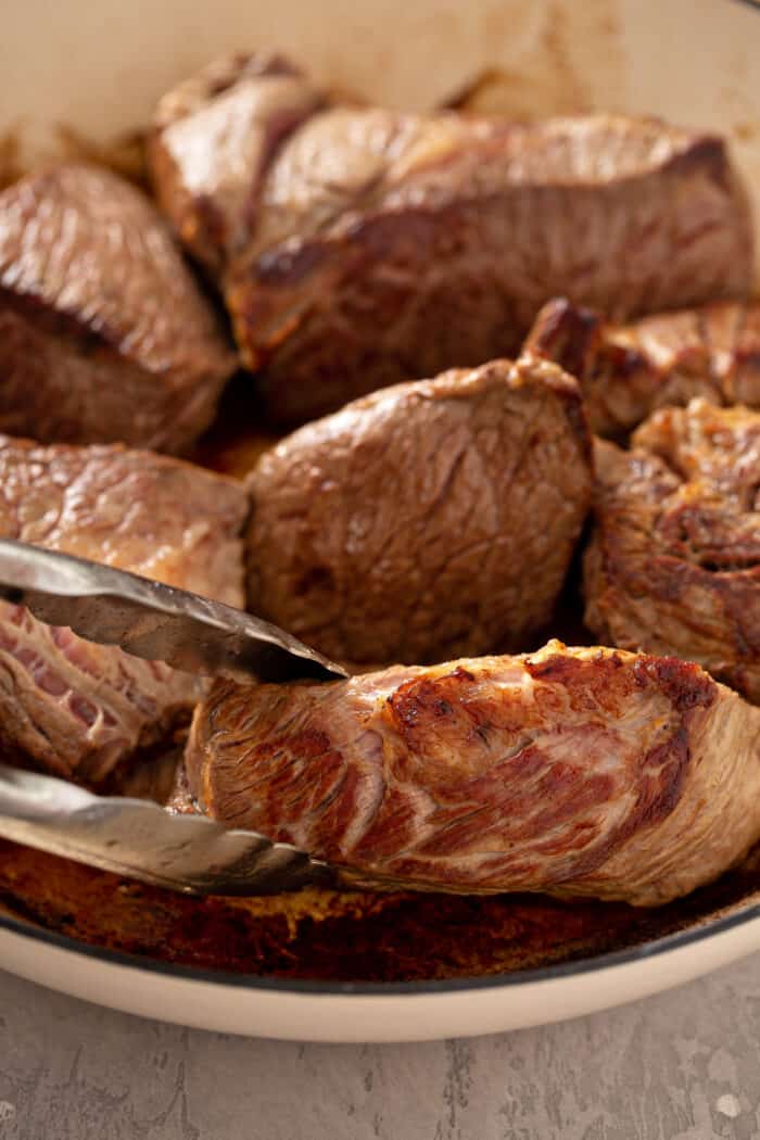 Tongs turning seared beef in a dutch oven to make chipotle barbacoa