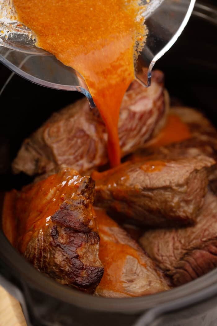 Bowl pouring chipotle sauce over seared beef in a slow cooker