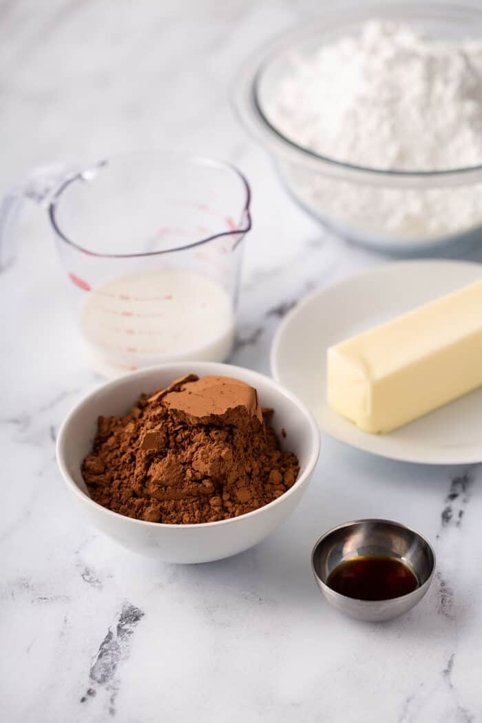 Ingredients for homemade chocolate frosting arranged in bowls on a marble counter