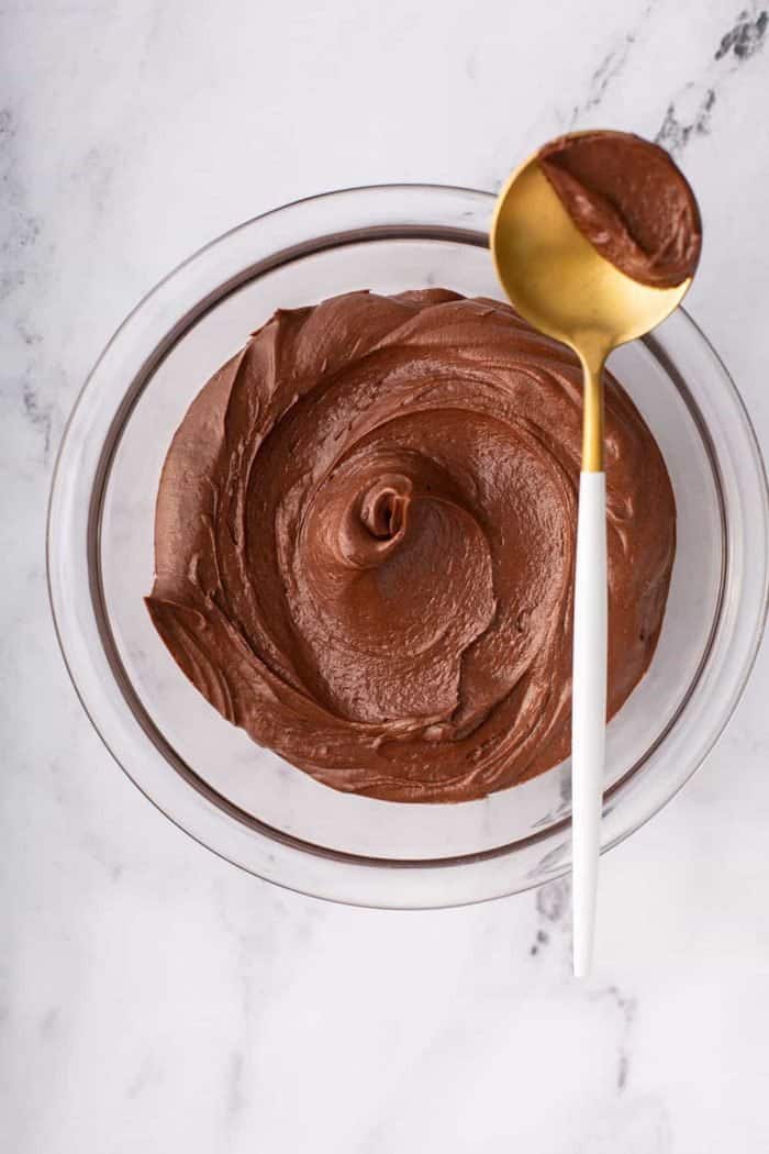Overhead view of homemade chocolate frosting in a glass bowl with a gold spoon balanced on the edge, holding a dollop of frosting