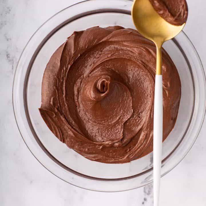Overhead view of homemade chocolate frosting in a glass bowl with a gold spoon balanced on the edge, holding a dollop of frosting