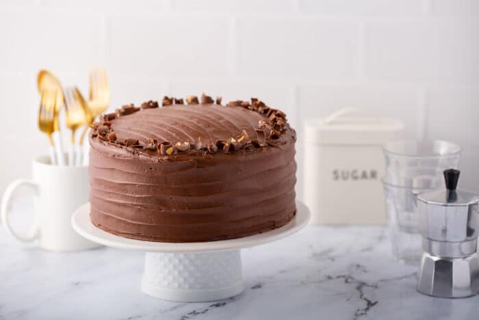 Chocolate-frosted Hershey's chocolate cake on a white cake stand on a marble countertop