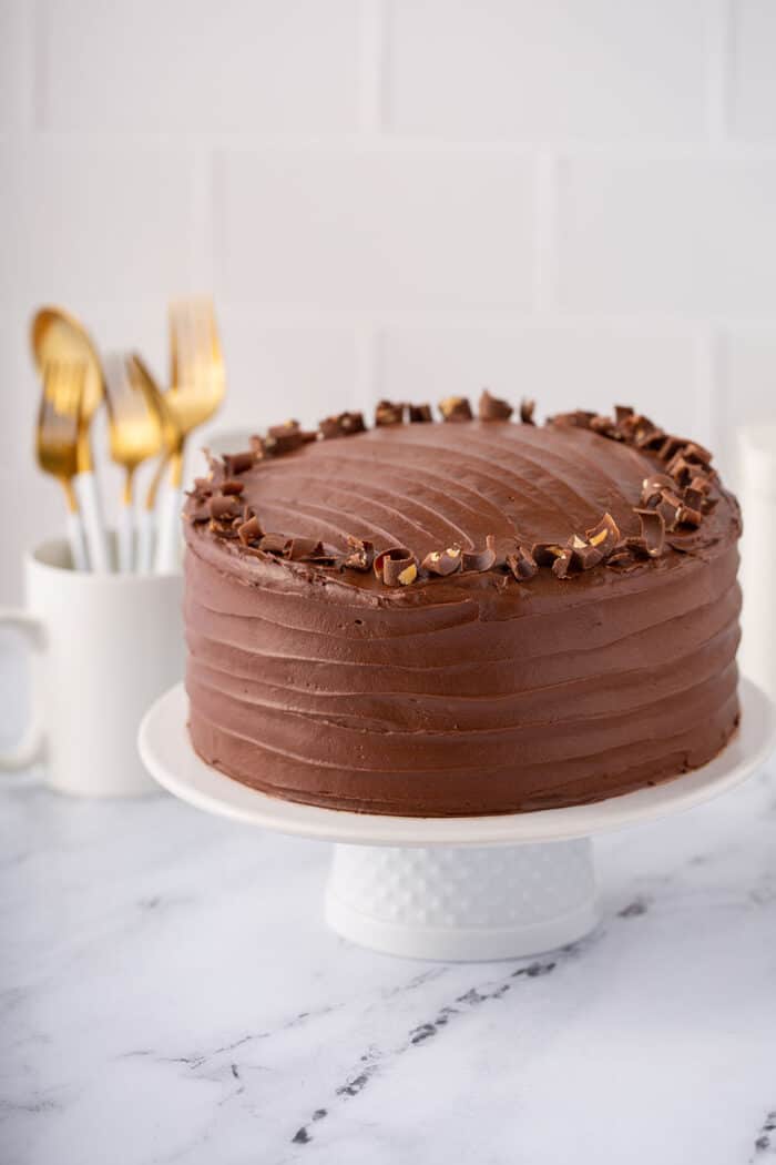 Hershey's chocolate cake frosted with chocolate frosting on a white cake plate with a mug holding forks in the background