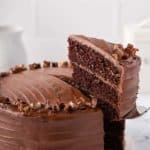 Cake server removing a slice of 3-layer Hershey's chocolate cake with chocolate frosting from a full cake on a white cake plate