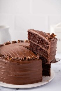 Cake server removing a slice of 3-layer Hershey's chocolate cake with chocolate frosting from a full cake on a white cake plate