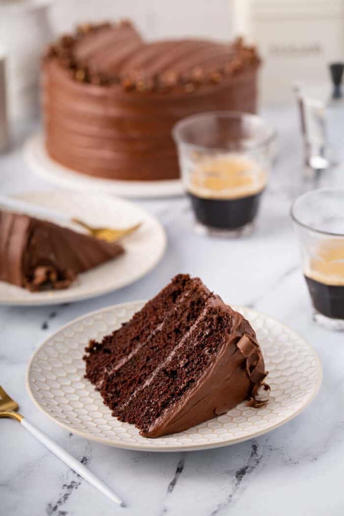 Slice of Hershey's Chocolate Cake with chocolate frosting on a white plate, with more cake and cups of espresso in the background