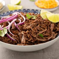 Chipotle barbacoa in a blue and white bowl topped with red onions, cilantro and lime wedges