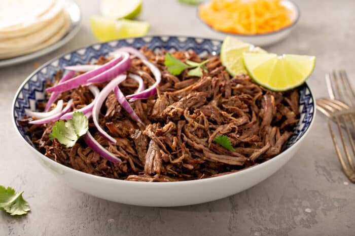 Chipotle barbacoa in a blue and white bowl topped with red onions, cilantro and lime wedges