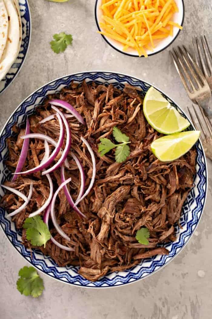 Overhead view of chipotle barbacoa in a blue and white bowl, toped with red onions, cilantro and lime wedges