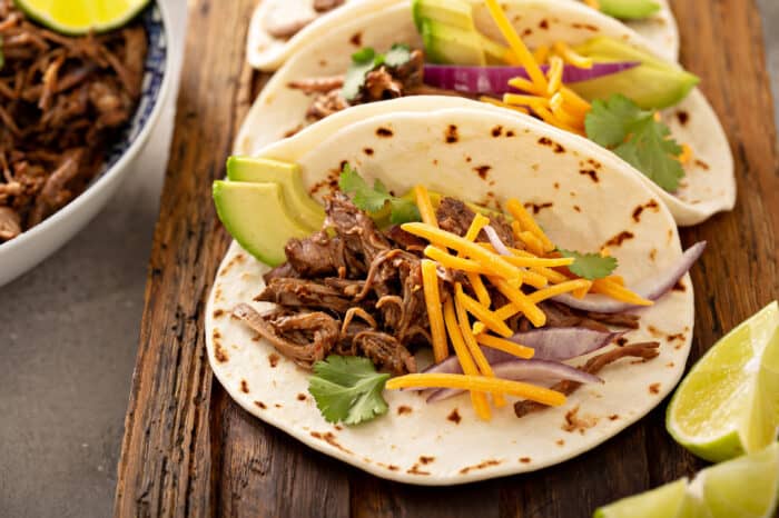 Close up of chipotle barbacoa tacos in flour tortillas, topped with cheese and sliced avocado, on a wooden board