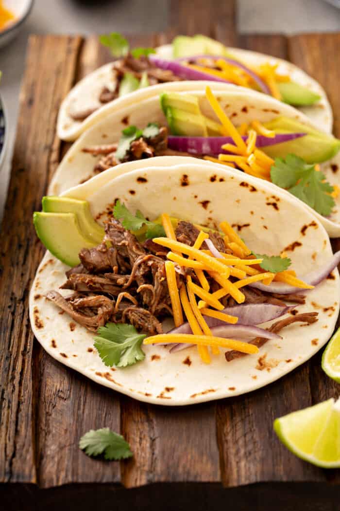 Three Chipotle barbacoa tacos on a wooden platter, topped with cheese and sliced avocado