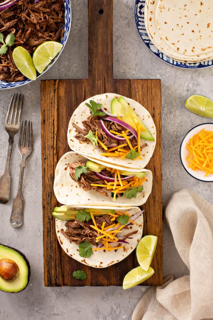 Vue de dessus de trois tacos de barbacoa au chipotle dans des tortillas de farine alignés sur une planche à découper en bois. planche à découper