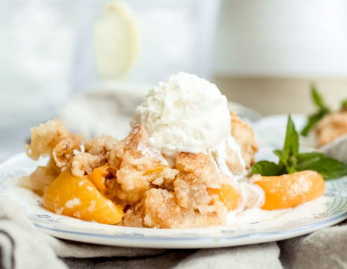 Close up of a serving of easy peach cobbler on a white plate topped with a scoop of melting vanilla ice cream