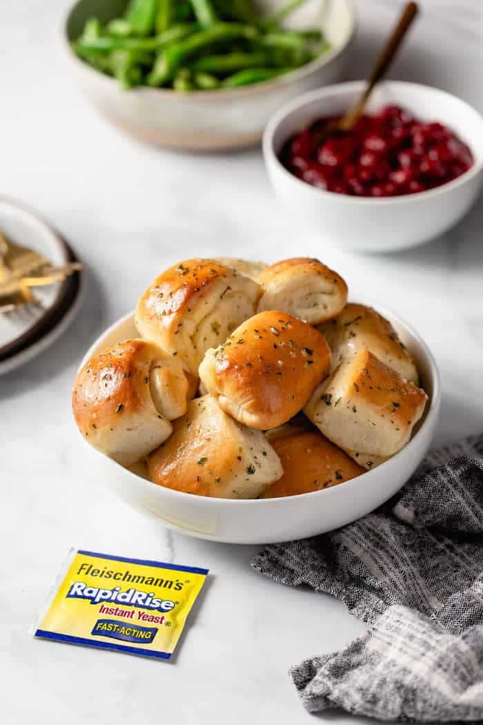 White bowl filled with garlic and herb parker house rolls on a counter surrounded by other holiday side dishes