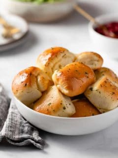 Close up of garlic and herb parker house rolls in a white bowl, set on a marble counter