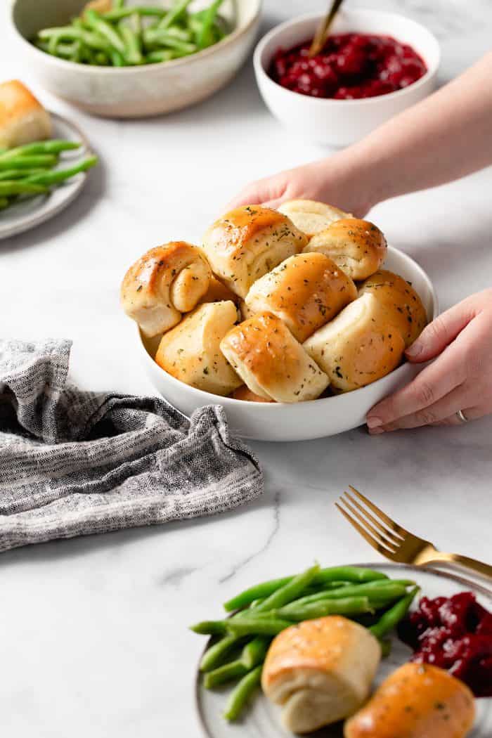 Hand placing a white bowl filled with garlic and herb parker house rolls on a marble counter filled with other holiday side dishes