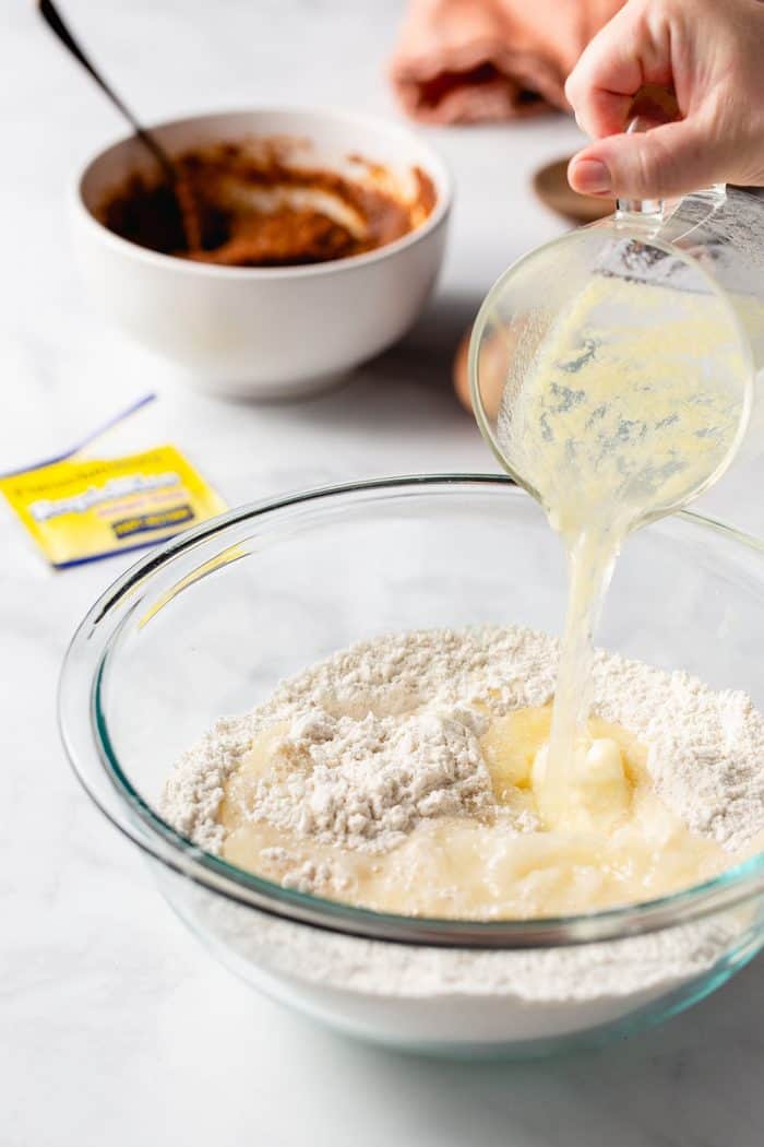 Hand pouring wet ingredients into dry ingredients in a glass mixing bowl for pumpkin spice cinnamon roll dough. A white bowl of pumpkin filling is in the background