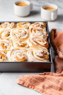 Baking pan of frosted pumpkin spice cinnamon rolls on a marble counter next to a dish towel