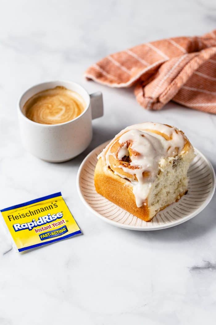 White plate with a pumpkin spice cinnamon roll next to a cup of coffee, with a package of yeast in the foreground