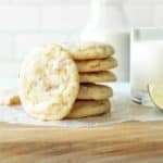 Chewy lime sugar cookies stacked on a parchment-covered cutting board next to a glass of milk