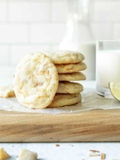 Chewy lime sugar cookies stacked on a parchment-covered cutting board next to a glass of milk