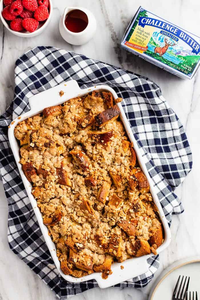 Overhead view of overnight french toast casserole in a white baking dish set on a blue plaid dish towel. A bowl of raspberries, a pitcher of syrup, and a package of butter are nearby.