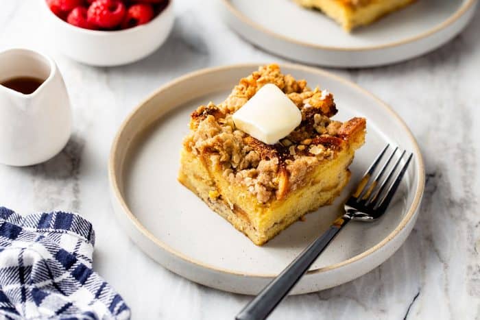 Slice of french toast casserole topped with a pat of butter next to a fork on a white plate. A pitcher of syrup and a bowl of raspberries are in the background.