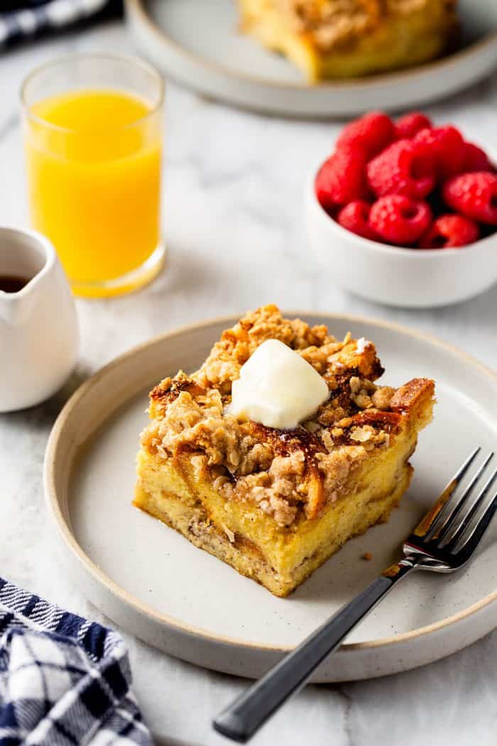 Slice of overnight french toast casserole topped with butter next to a fork on a white plate. Orange juice and fresh raspberries are in the background