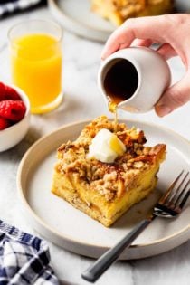 Hand drizzling maple syrup over a slice of buttered overnight french toast casserole on a white plate. A glass of orange juice is in the background