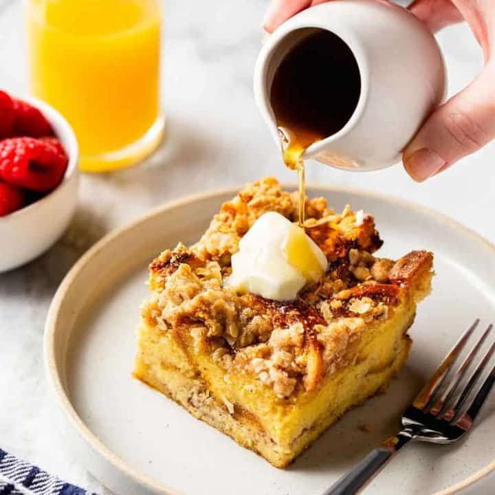 Hand drizzling maple syrup over a slice of buttered overnight french toast casserole on a white plate. A glass of orange juice is in the background