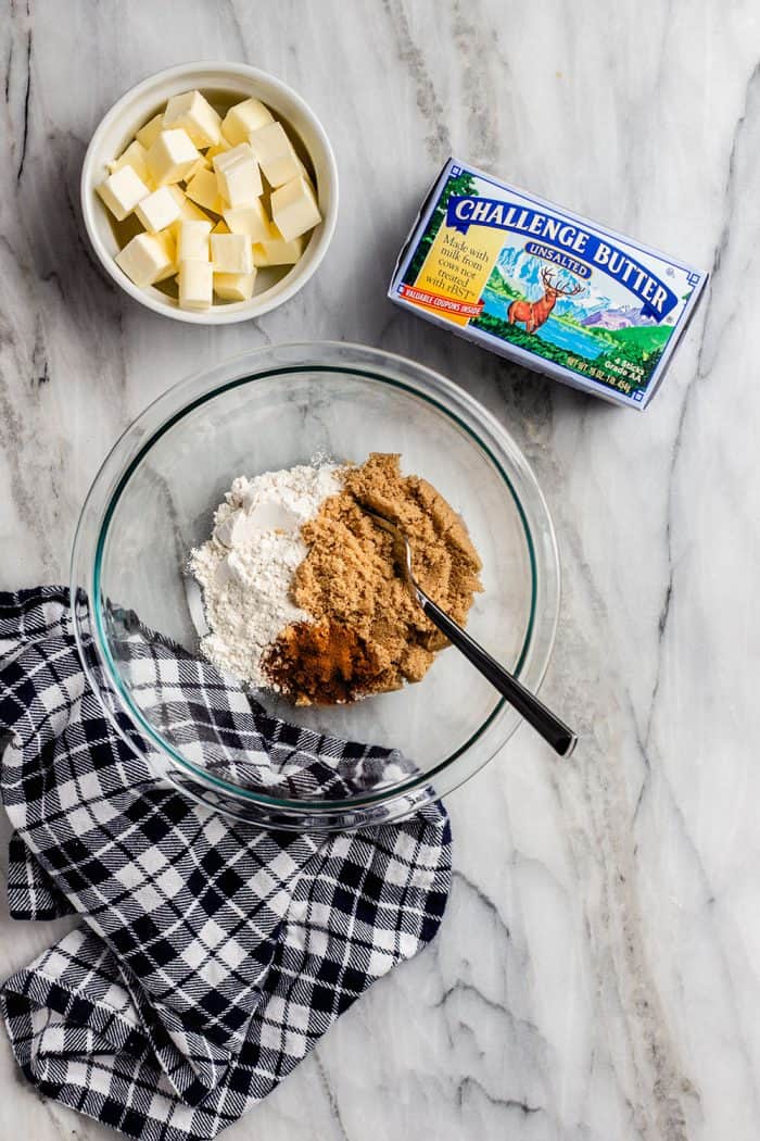 Overhead view of dry ingredients for french toast casserole streusel topping in a glass bowl on a marble countertop. A bowl of butter and butter package is set nearby.