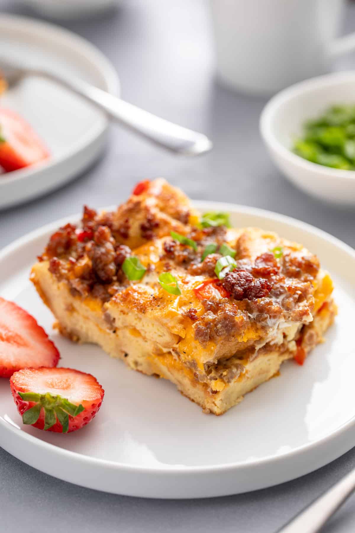 Close up of a slice of make-ahead breakfast casserole next to a halved strawberry on a white plate.