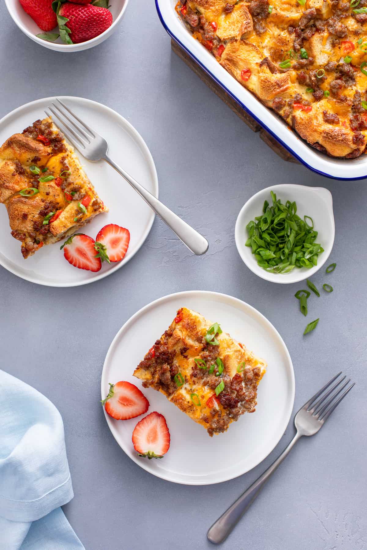 Overhead view of two white plates, each holding a halved fresh strawberry and a slice of make-ahead breakfast casserole.