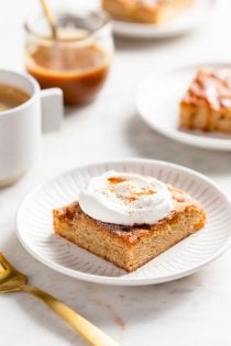 Apple bar topped with whipped cream on a white plate with caramel, coffee, and more plated apple bars in the background