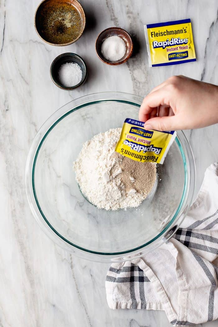 Hand pouring a package of yeast into a bowl of flour on a marble surface