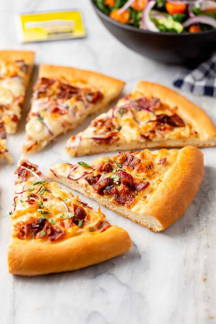 Slices of chicken bacon ranch pizza on a marble surface with a bowl of salad in the background